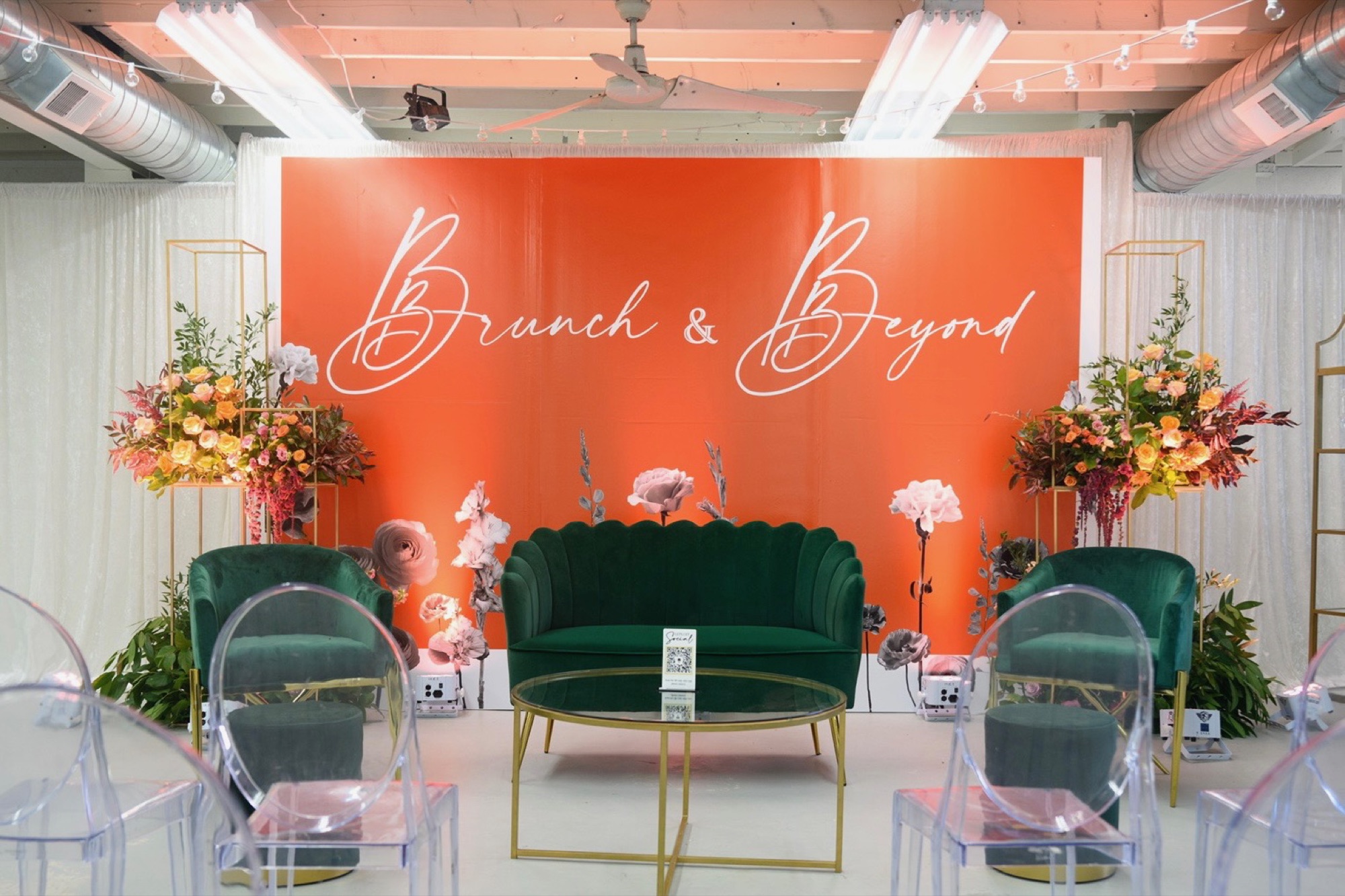 Brunch & Beyond lounge set with an orange script backdrop, green velvet loveseat and chairs, gold-framed florals, and clear ghost chairs