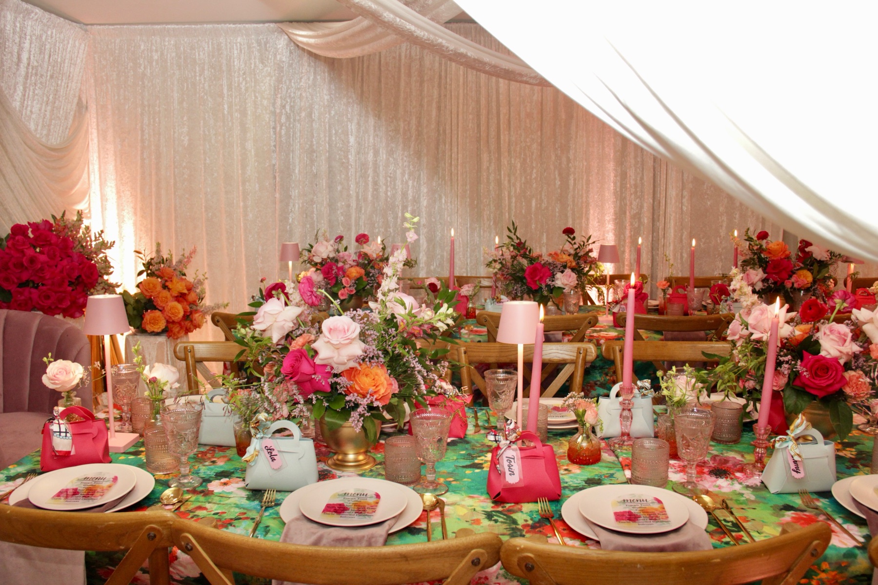 Long banquet table with pink and coral centerpieces, taper candles, blush lamps, gift-bag place settings, and a painted floral tablecloth