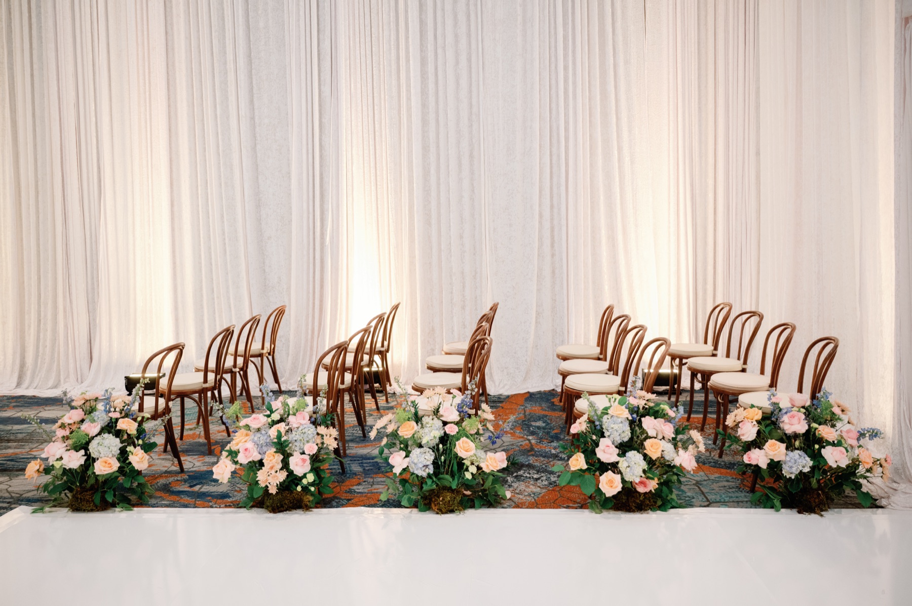Rows of bentwood ceremony chairs in side profile, lined with pastel rose and hydrangea arrangements before ivory drapery